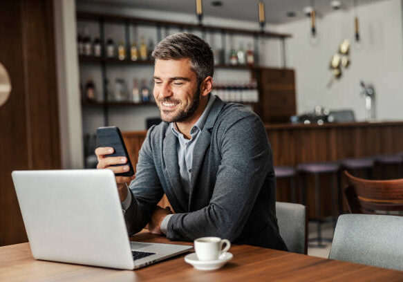 A happy businessman is sitting in a coffee shop and checking on his bank account on the mobile. There is a laptop on a table. A man using the phone for e-banking